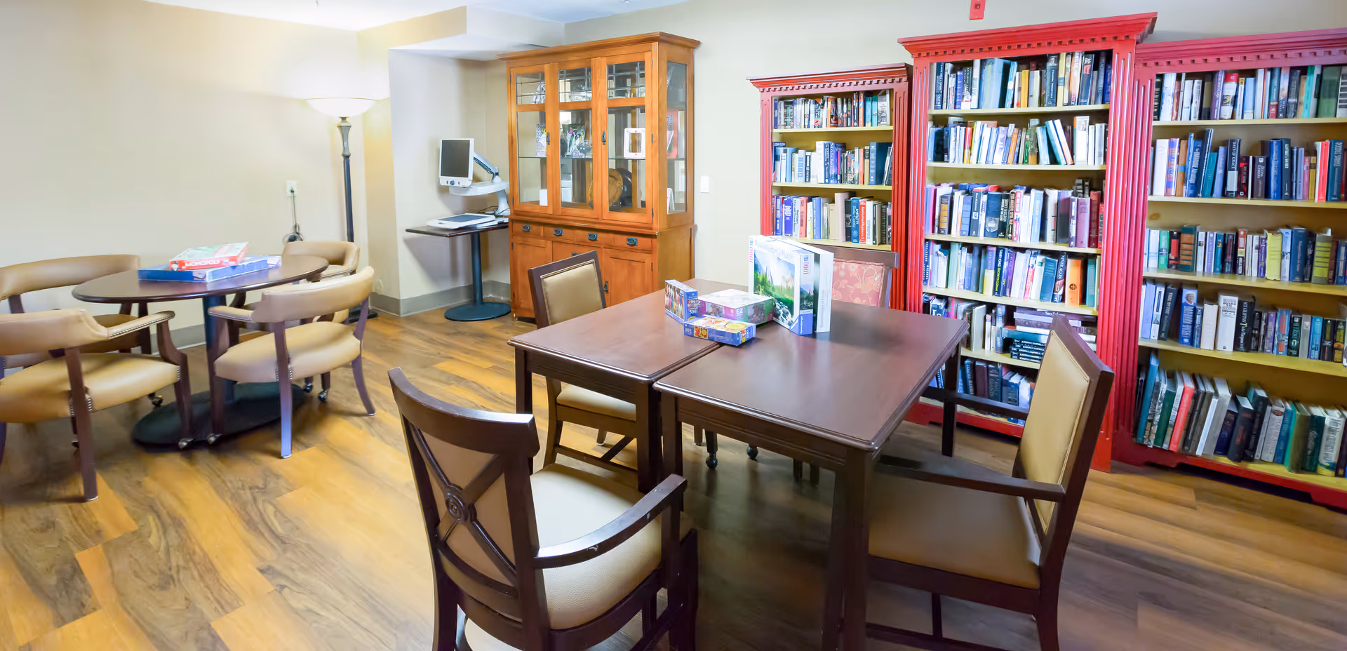 A cozy room with wooden flooring featuring two sets of tables and chairs. One set has a rectangular table with four chairs and several puzzle boxes on top. The other set has a round table with four chairs and board games on it. There are three large bookshelves filled with books along one wall, two of which are painted red. A wooden cabinet with glass doors and a computer workstation with a monitor are also visible. A tall floor lamp provides lighting in the corner.