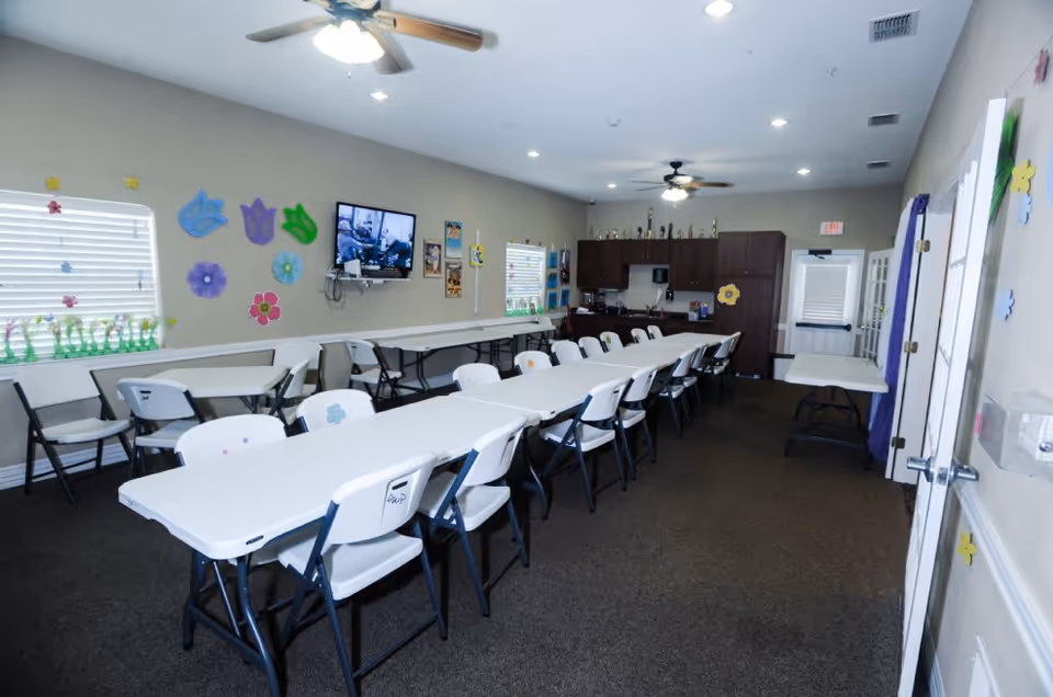 A long room with white folding tables and chairs arranged in rows, decorated with colorful paper flowers on the walls. There are two ceiling fans with lights, a wall-mounted TV, and a kitchenette area with cabinets and a coffee maker at the far end. Windows with blinds let in natural light.