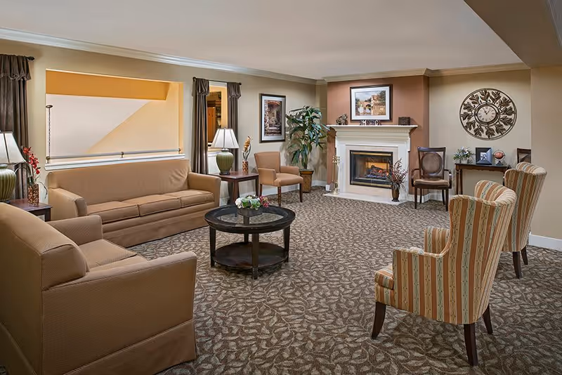 A cozy senior living facility common area with beige and brown tones, featuring a fireplace with a mantel decorated with a framed picture and small ornaments. The room has a patterned carpet, two beige sofas, two striped armchairs, a round dark wood coffee table with a floral centerpiece, side tables with lamps, a large wall clock, framed artwork, and a potted plant near the fireplace.