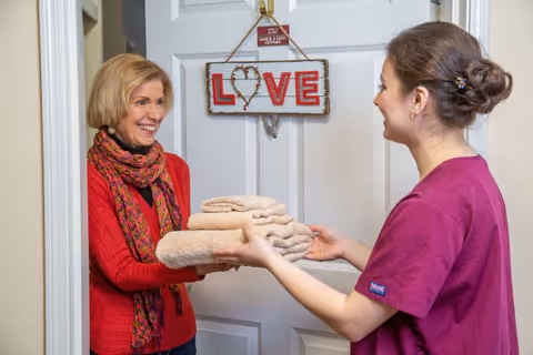 A smiling elderly woman in a red sweater and scarf receives a stack of folded towels from a caregiver wearing a purple uniform in front of a white door with a decorative sign that says 'LOVE'.