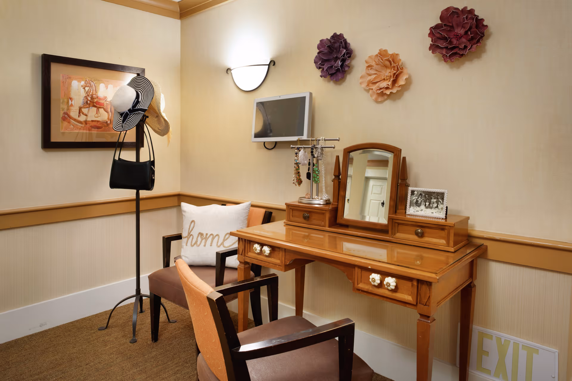 Cozy interior dressing area with a wooden vanity and mirror, two chairs, a hat stand, decorative wall flowers and a small wall-mounted TV.
