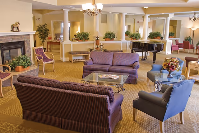 Communal lounge with multiple sofas and armchairs arranged around a coffee table, a fireplace, plants and a grand piano.