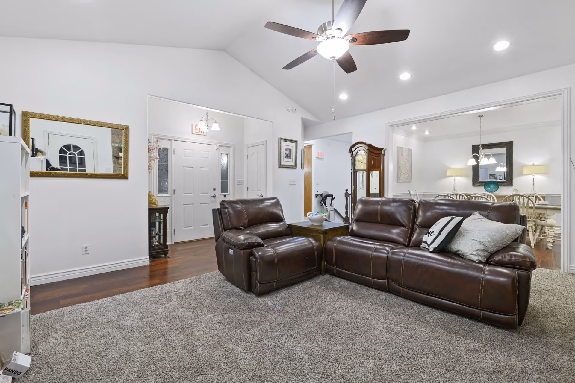 A cozy living room with a brown leather reclining sofa and chair on a beige carpet. The room has white walls, a ceiling fan with lights, and recessed lighting. A large mirror hangs on the left wall above a white bookshelf. In the background, there is a dining room with a white dining table and chairs, illuminated by two table lamps and a hanging light fixture. The front door with sidelights is visible, along with a grandfather clock near the entrance to the dining room.