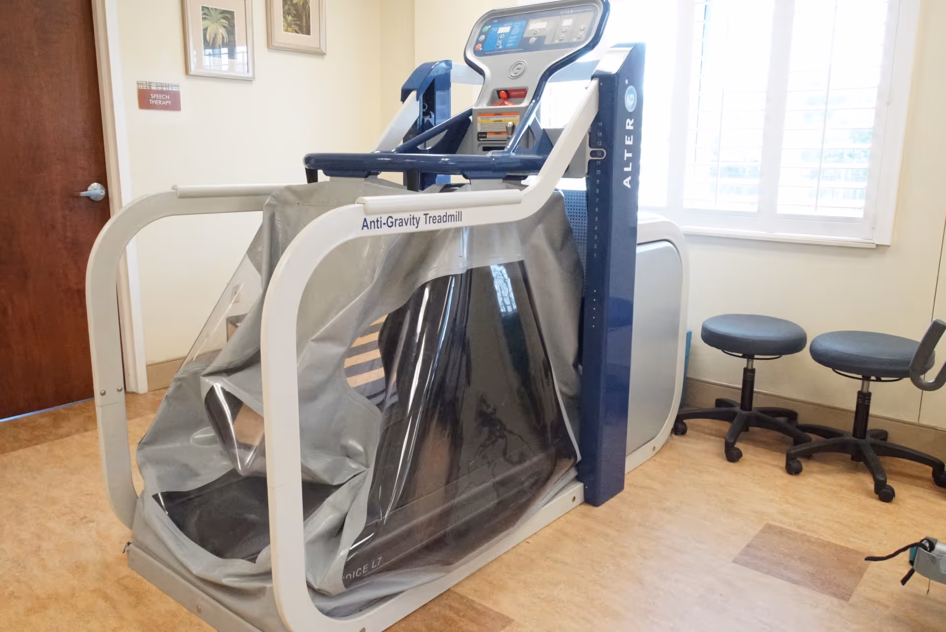 An anti-gravity treadmill machine in a therapy room with two black stools, a window with white shutters, and a door with a sign that reads 'Speech Therapy'. The room has light-colored walls and a wood-patterned floor.