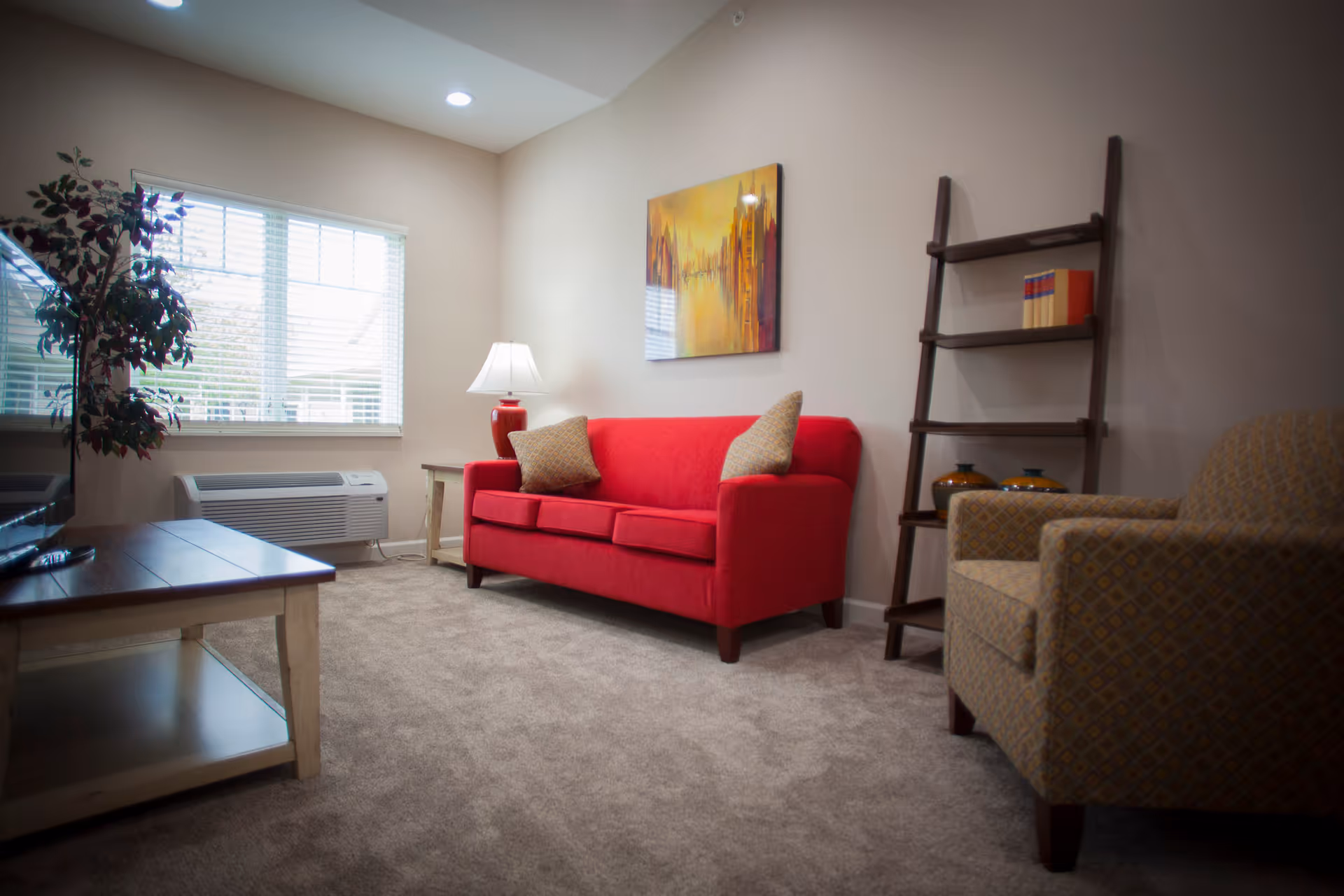 A cozy living room with a red sofa adorned with two beige cushions, a beige patterned armchair, a wooden coffee table, a tall wooden ladder shelf with decorative items, a side table with a red lamp, a large window with blinds, and a colorful abstract painting on the wall.
