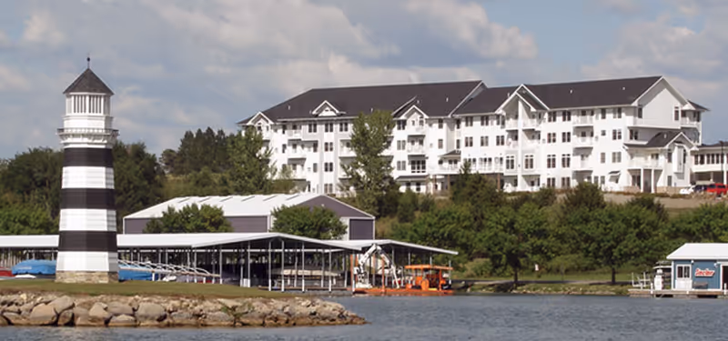 A large white multi-story building with balconies situated on a hillside overlooking a body of water. In the foreground, there is a black and white striped lighthouse, a covered boat dock with boats, and a small building near the water's edge. Trees and greenery surround the area under a partly cloudy sky.