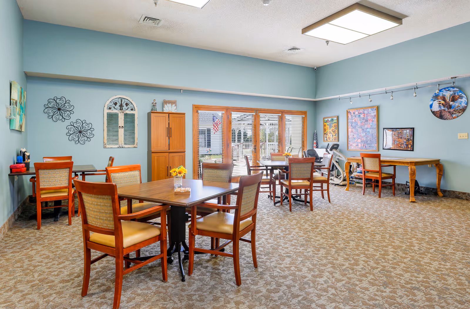 A spacious, well-lit common area with several wooden tables and chairs arranged for seating. The walls are painted light blue and decorated with various framed artworks and decorative items. Large windows with wooden blinds allow natural light to enter the room. The carpet has a patterned design, and there is an exercise bike near the back wall.