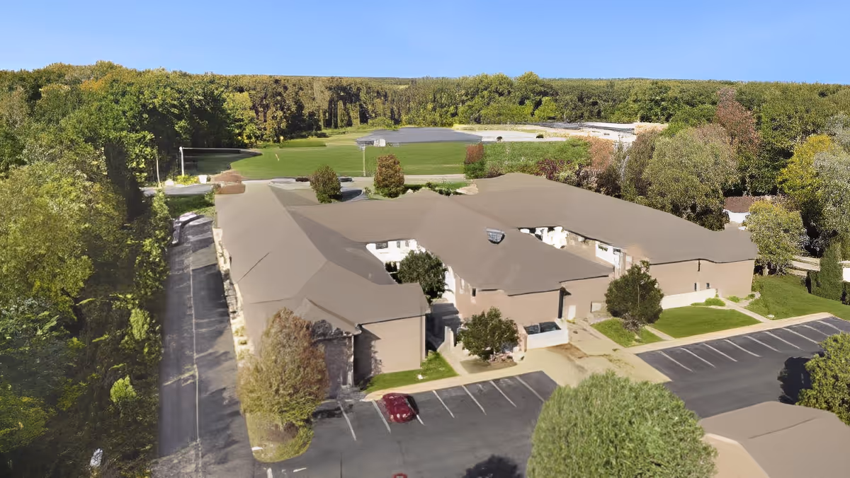 Aerial view of a senior living facility building surrounded by trees and greenery, with parking lots in the foreground and a clear blue sky above.