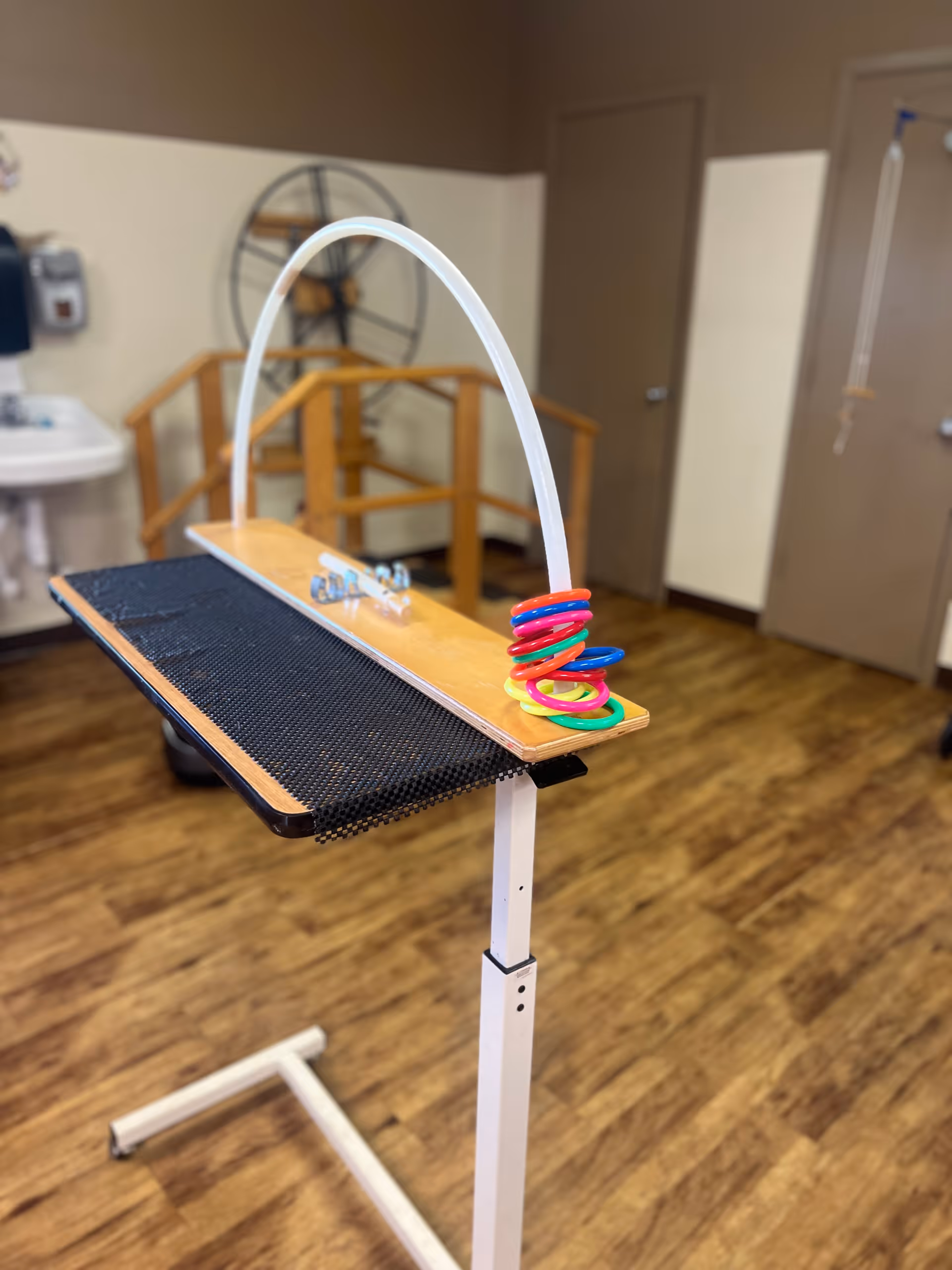A therapy or rehabilitation room with a wooden and metal exercise device featuring colorful plastic rings on a white stand. The room has wood flooring, beige and brown walls, a sink, and a wooden handrail in the background.