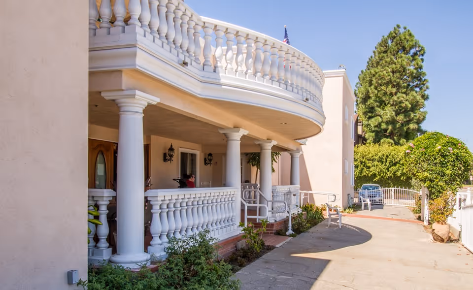 Exterior view of a senior living facility with a curved balcony supported by white columns, a walkway with plants along the side, and a gated entrance. There is a person sitting on the porch under the balcony.