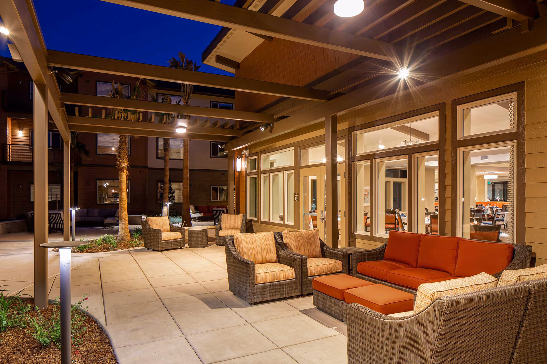 Outdoor patio area at night with wicker furniture including chairs and a sofa with orange cushions, under a pergola structure. The patio is adjacent to a building with large windows showing an indoor dining area with tables and chairs. Palm trees and soft lighting enhance the ambiance.