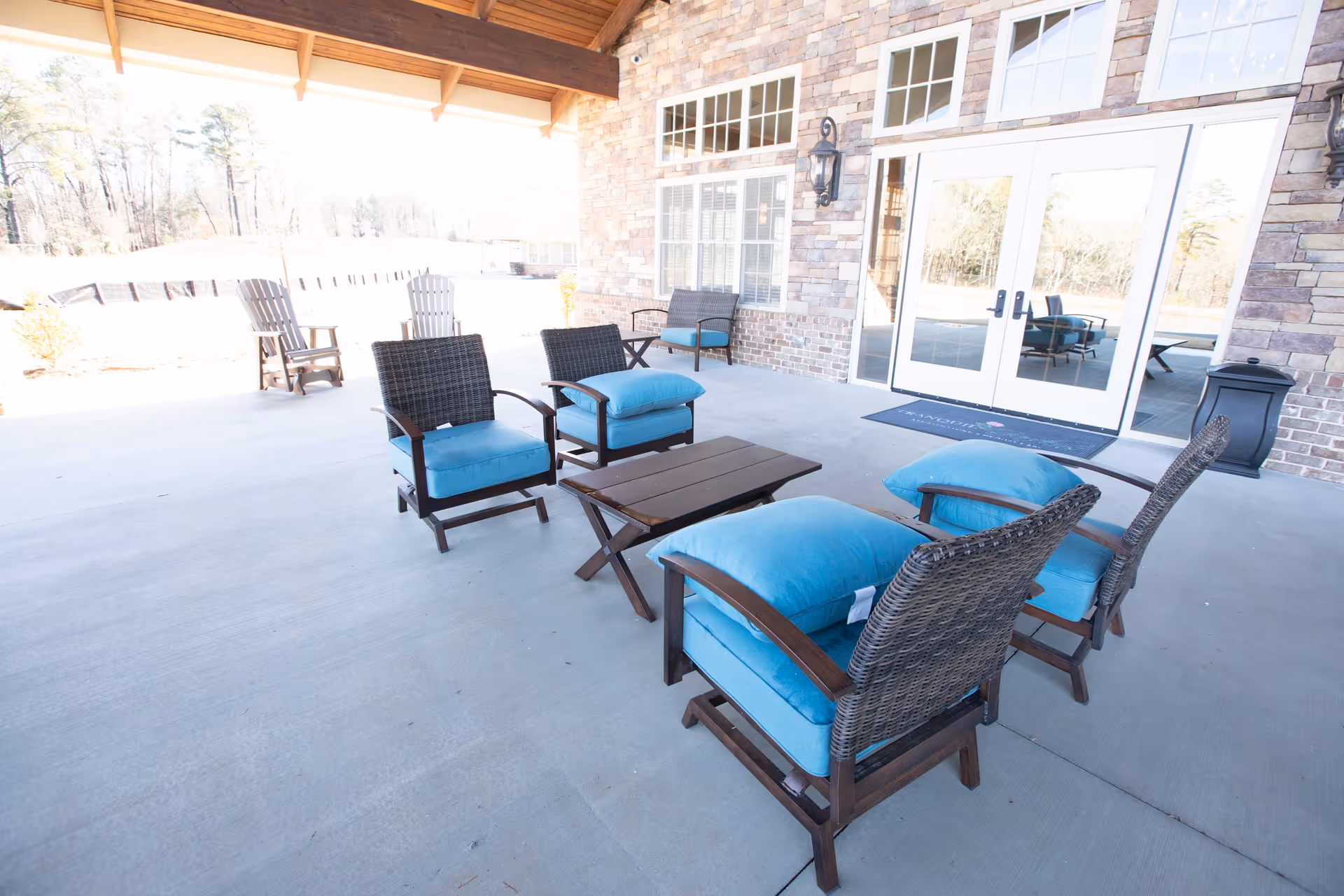 Outdoor covered patio area with wicker chairs featuring blue cushions arranged around a wooden coffee table. The patio has a concrete floor and a wooden ceiling. There are large windows and double glass doors leading inside a brick building. Additional seating is visible in the background along with trees and open outdoor space.