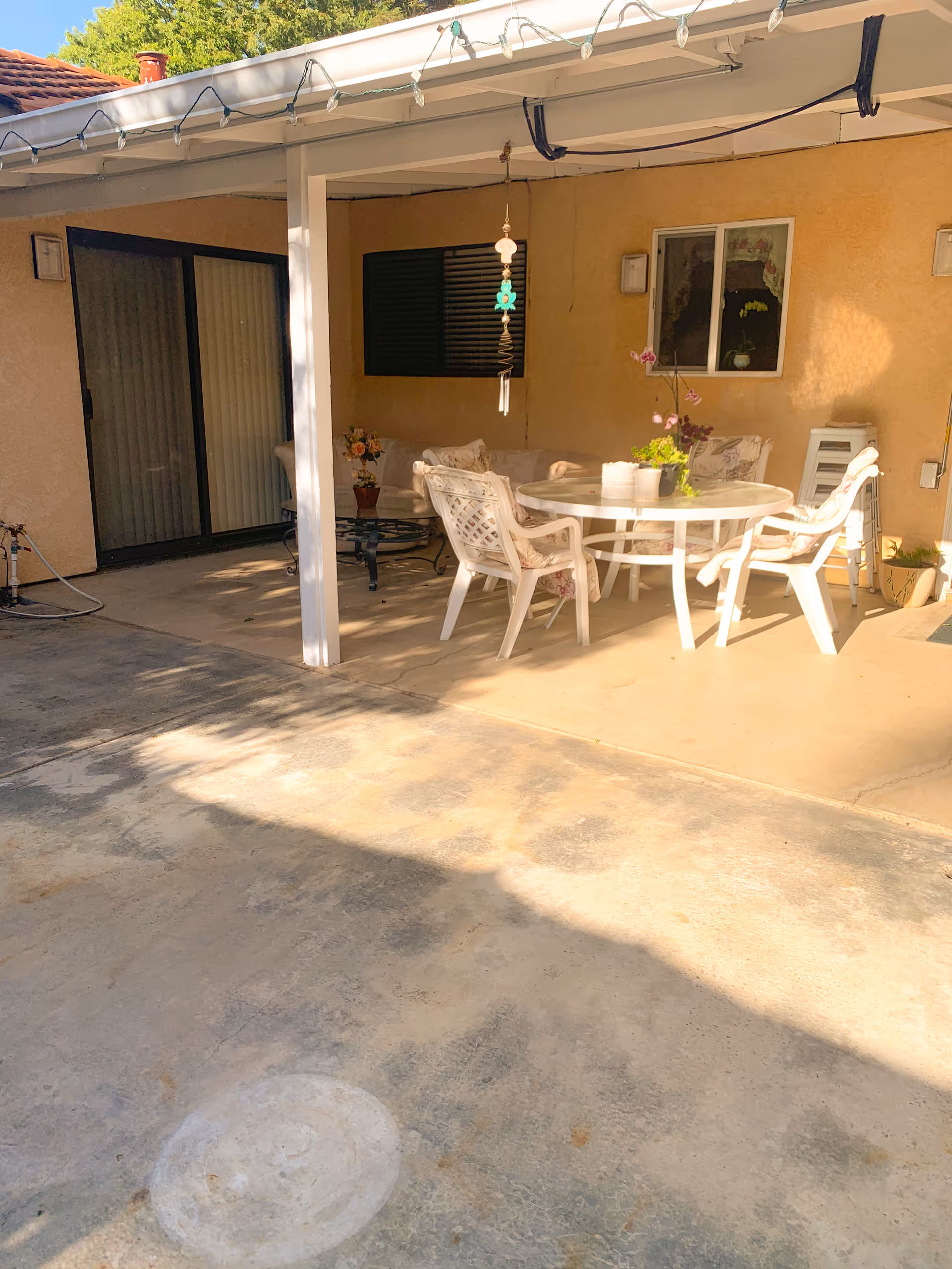Outdoor covered patio area with a round white table and four white chairs with cushions. There are potted plants on the table and nearby, a hanging decoration, sliding glass doors, and a window on the beige wall of the building.