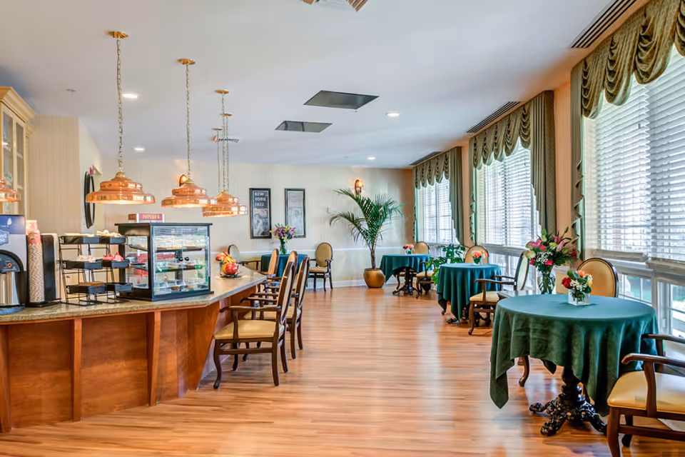 A bright dining area with wooden floors and large windows covered with green valances. Several round tables with green tablecloths and floral centerpieces are arranged along the windows. On the left side, there is a counter with copper pendant lights hanging above, chairs lined up, and a display case with snacks. The walls are decorated with framed pictures and a large potted plant is placed in the corner.