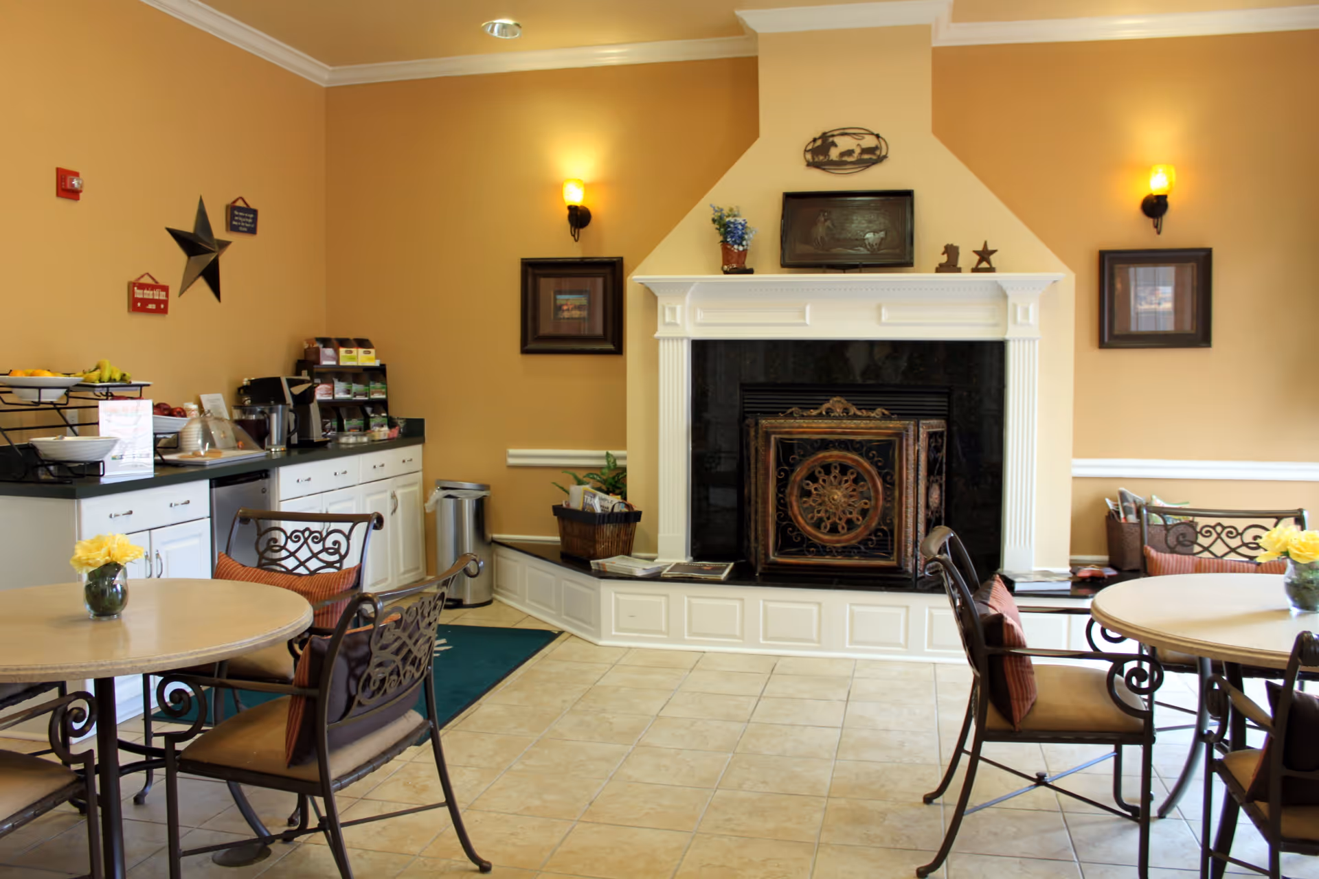 A cozy common area with beige walls and tiled floor featuring a white fireplace with decorative black and gold screen. Two round tables with wrought iron chairs and cushions are placed in the room. A counter with snacks, coffee machine, and condiments is against one wall. The room is softly lit by wall sconces and decorated with framed pictures and small decor items.