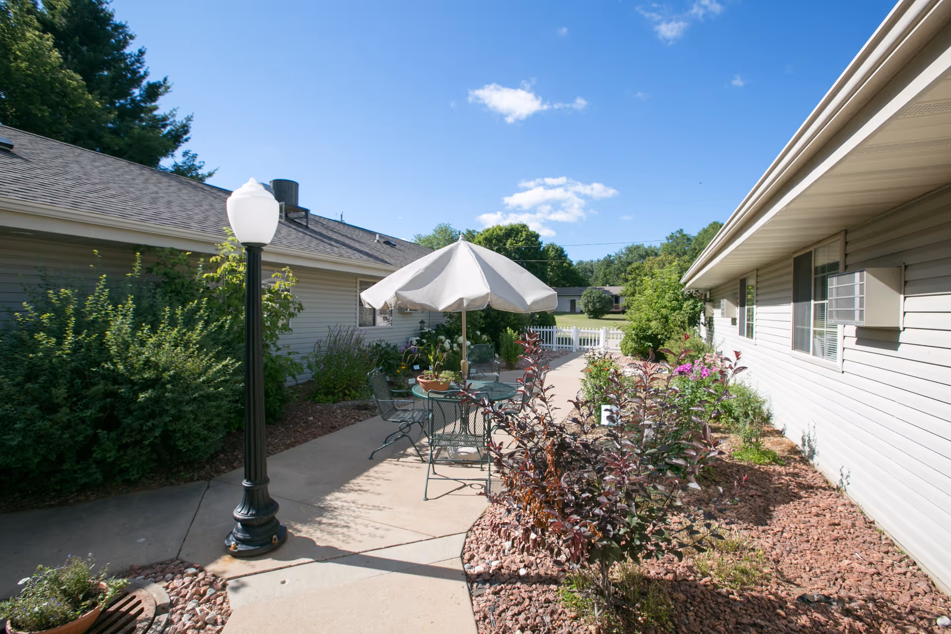 Outdoor patio area between two single-story buildings with beige siding, featuring a round metal table with chairs and a large white umbrella. The patio is surrounded by landscaped garden beds with various plants and shrubs, under a clear blue sky with a few clouds.