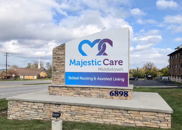 Outdoor stone sign for Majestic Care Middletown Skilled Nursing & Assisted Living, located on a grassy area near a road with buildings and trees in the background under a partly cloudy sky.
