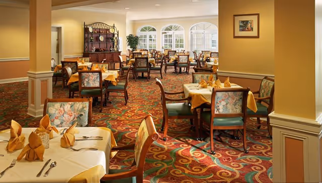 A spacious dining room with multiple tables covered in yellow tablecloths and neatly folded napkins. The chairs have floral upholstery and wooden frames. The room features large windows allowing natural light to fill the space, a patterned carpet with red, green, and yellow swirls, and a wooden cabinet against the far wall.