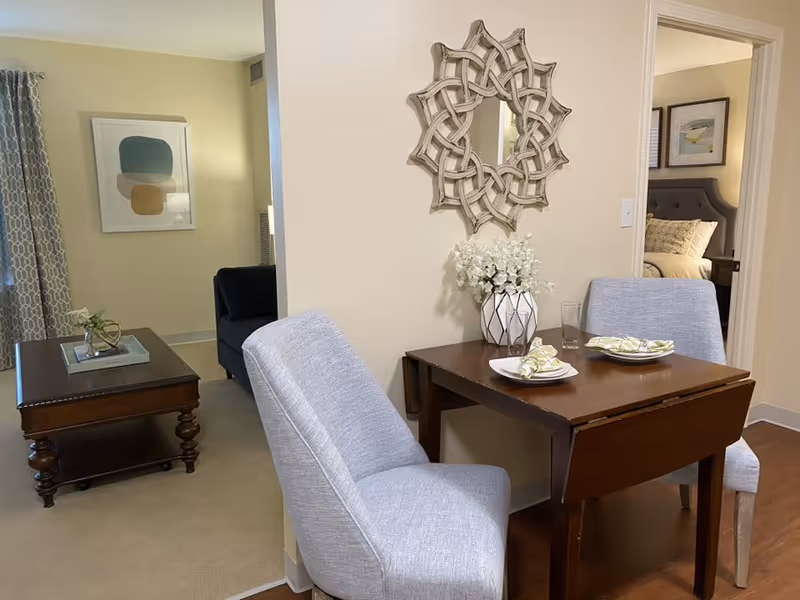 A cozy interior space featuring a small wooden dining table set for two with plates, napkins, and a glass. Two light gray upholstered chairs are positioned at the table. A decorative mirror with a woven design hangs on the wall above a vase with white flowers on the table. In the background, there is a living area with a dark sofa, a wooden coffee table with a small plant, and a framed abstract artwork on the wall. A bedroom with a bed and framed pictures is visible through an open doorway.