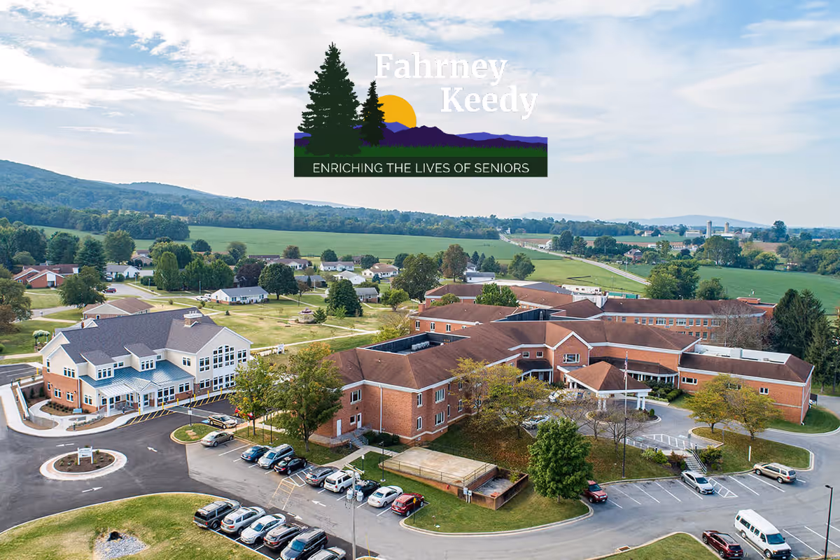 Aerial view of Fahrney-Keedy Senior Living Community showing multiple brick buildings with red roofs surrounded by parking lots, trees, and green fields under a partly cloudy sky. The facility is set in a rural landscape with hills in the background.
