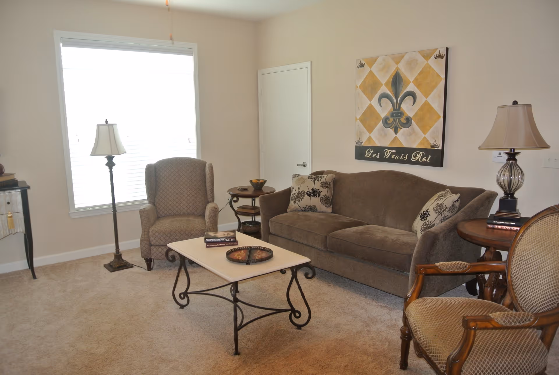 A cozy living room with a brown sofa adorned with patterned cushions, a beige armchair, and a wooden chair with a cushioned seat. There is a rectangular coffee table with a decorative tray and books on it. Two table lamps and a floor lamp provide lighting. A large window with blinds lets in natural light. A decorative wall art piece with a fleur-de-lis and the text 'Les Trois Roi' hangs above the sofa.