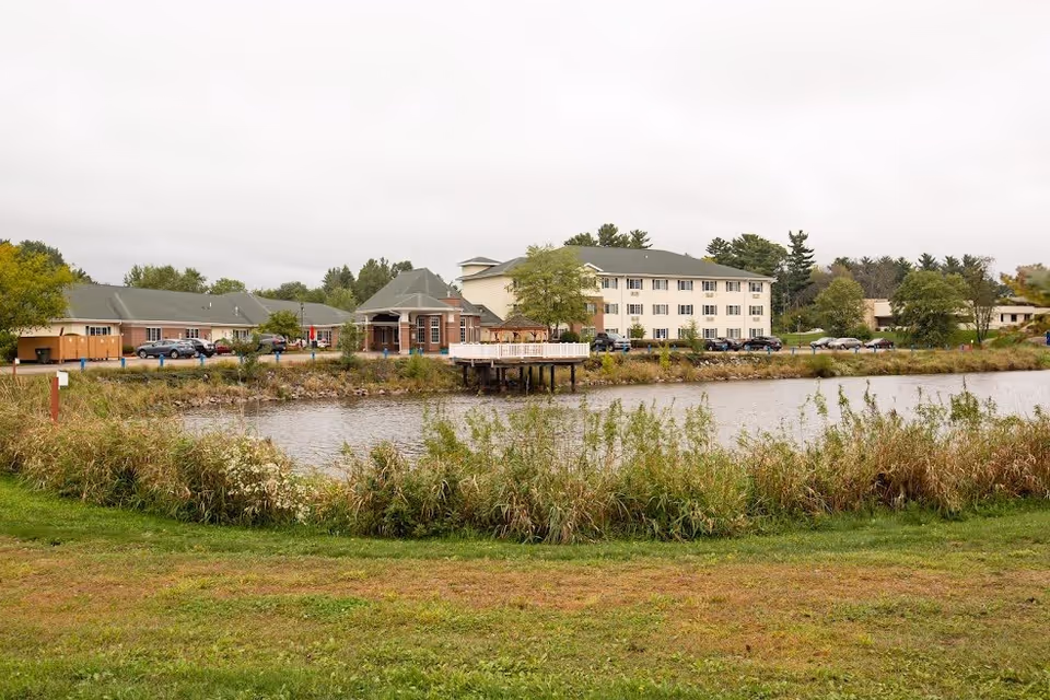 Exterior view of a senior living complex and adjacent buildings across a pond with grassy foreground.