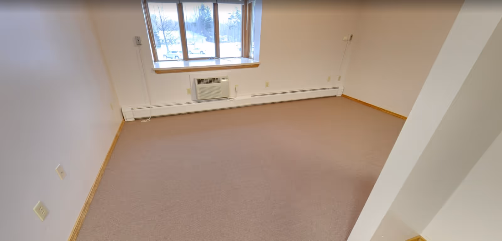 Empty room with beige carpet, white walls, a window with a view of snow outside, and a wall-mounted air conditioning unit below the window.
