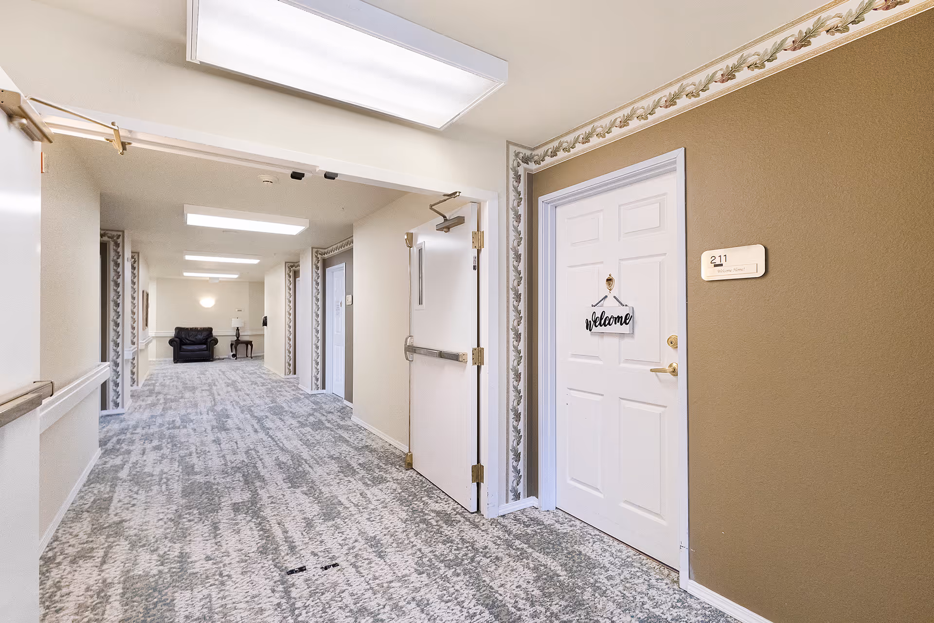 A well-lit hallway in a senior living facility with patterned carpet and beige walls. There is a white door on the right with a 'welcome' sign and a room number plaque labeled 211. At the end of the hallway, there is a black armchair and a small table with a lamp. The hallway has handrails along the walls and overhead fluorescent lighting.
