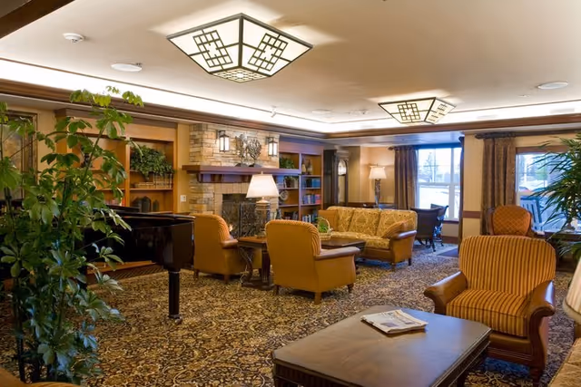A cozy and well-lit living room in The Stafford Retirement Community featuring a patterned carpet, a stone fireplace with a decorative metal piece above it, wooden bookshelves filled with books and plants, a grand piano, several upholstered armchairs and sofas, wooden coffee tables, and large windows with curtains allowing natural light to enter.