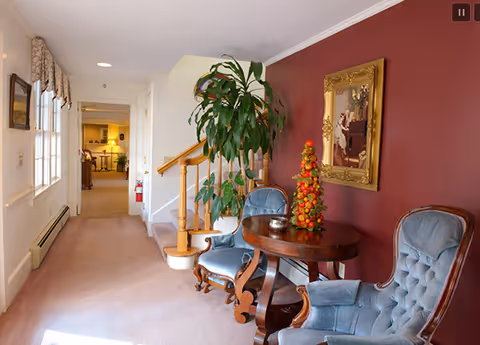A cozy interior hallway with beige carpet and white walls, featuring a wooden staircase with a handrail. Two blue upholstered armchairs and a round wooden table with a decorative centerpiece are placed against a maroon accent wall adorned with a framed painting. A large green plant is positioned near the staircase, and natural light enters through windows on the left side.