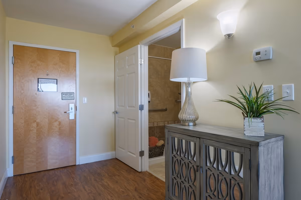 Interior view of a senior living facility room entrance area with a wooden door on the left, an open white door revealing a bathroom with tiled walls and a grab bar, and a decorative cabinet on the right topped with a lamp and a potted plant.