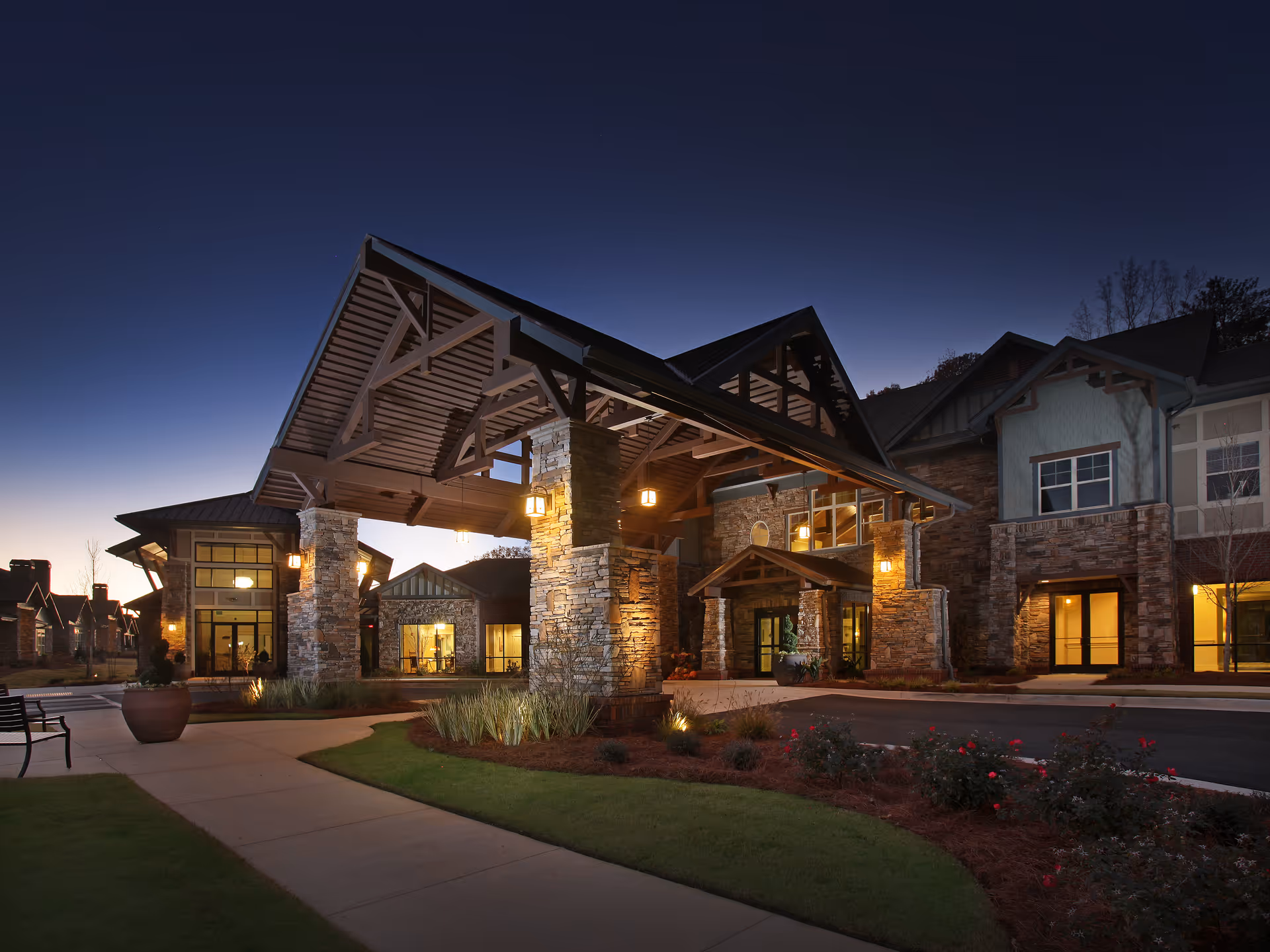 Exterior view of a senior living facility at dusk with stone pillars, a large covered entrance, warm lighting, landscaped greenery, and a clear sky.