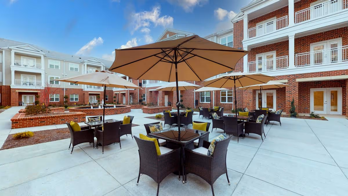 Outdoor courtyard with patio tables, umbrellas, and wicker chairs in front of a multi-story brick senior living building.