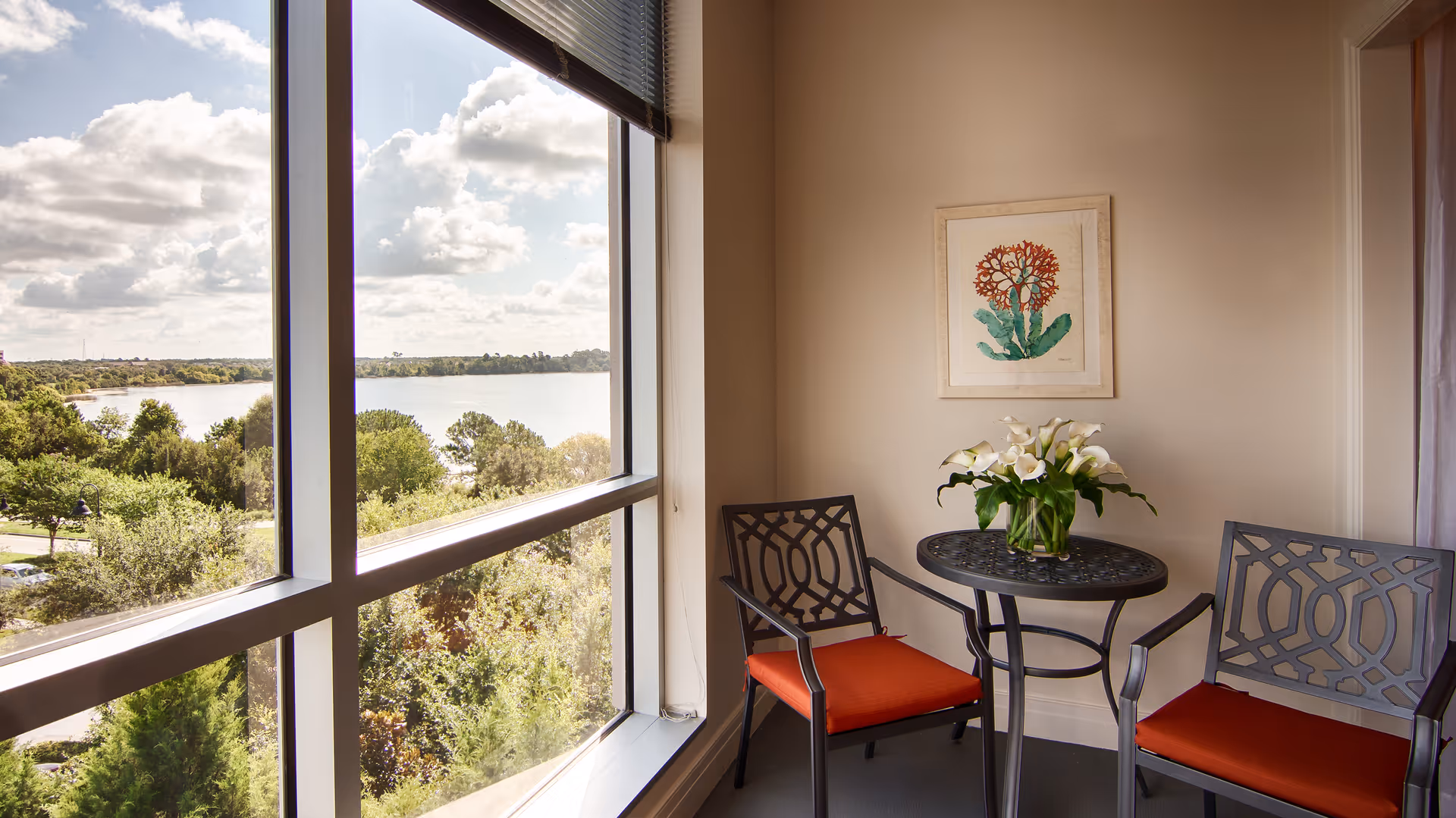 Small seating area with two metal chairs with orange cushions and a round table holding a vase of white flowers beside a large window overlooking a lake.