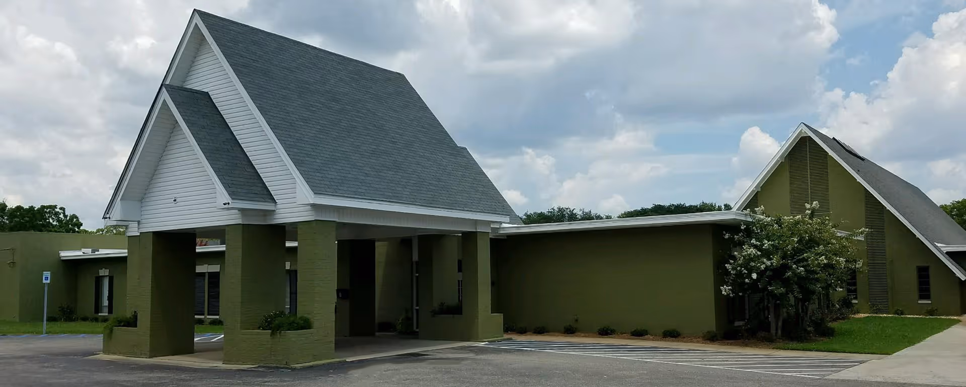 Exterior view of a senior living facility building with green walls and a large covered entrance with a steep gray roof. The sky is partly cloudy and there is a parking area in front of the building.
