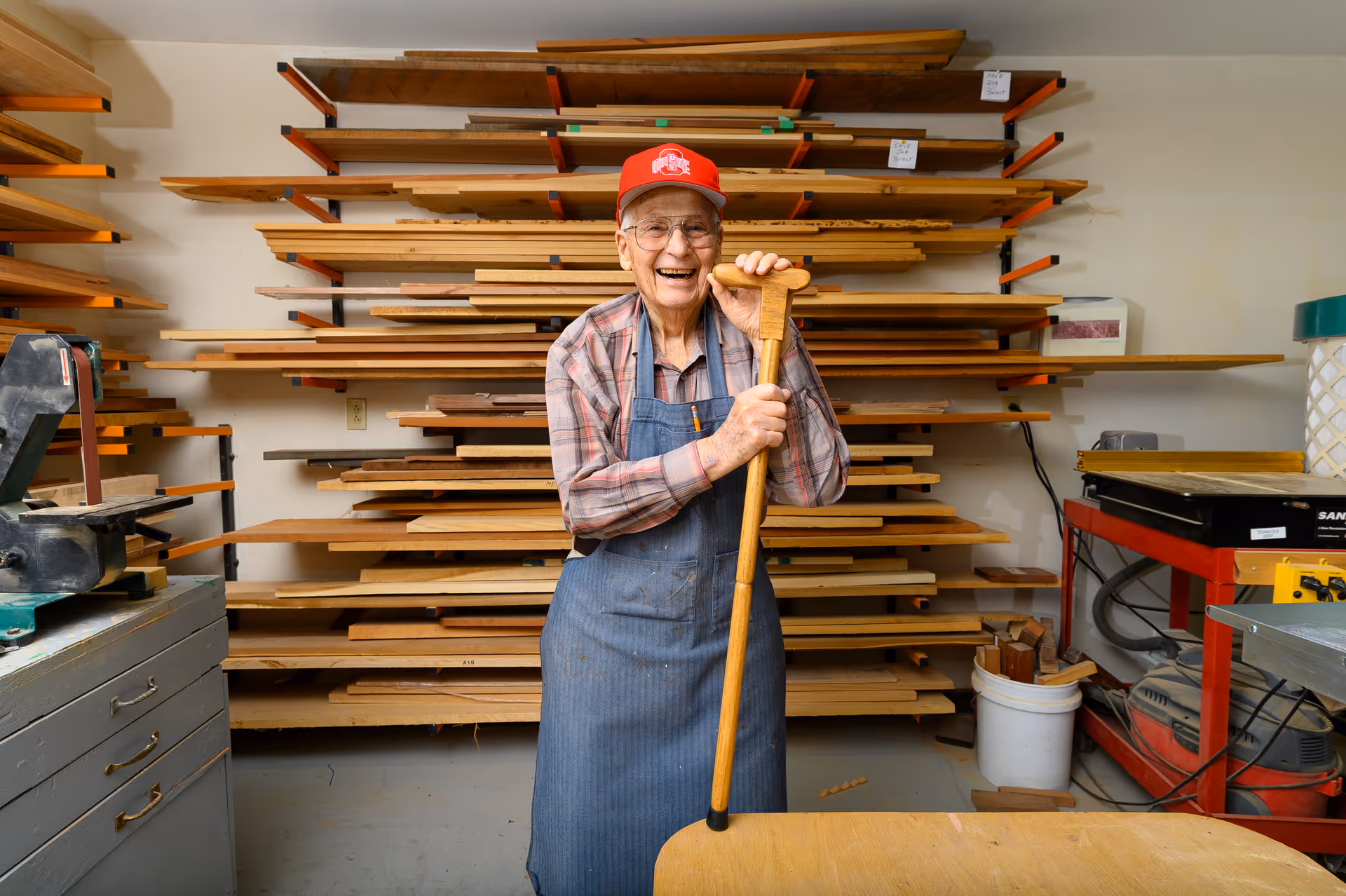 An elderly man wearing a red cap, glasses, plaid shirt, and blue apron stands smiling in a woodworking shop. He is holding a wooden cane and is surrounded by shelves filled with various wooden planks and woodworking tools.