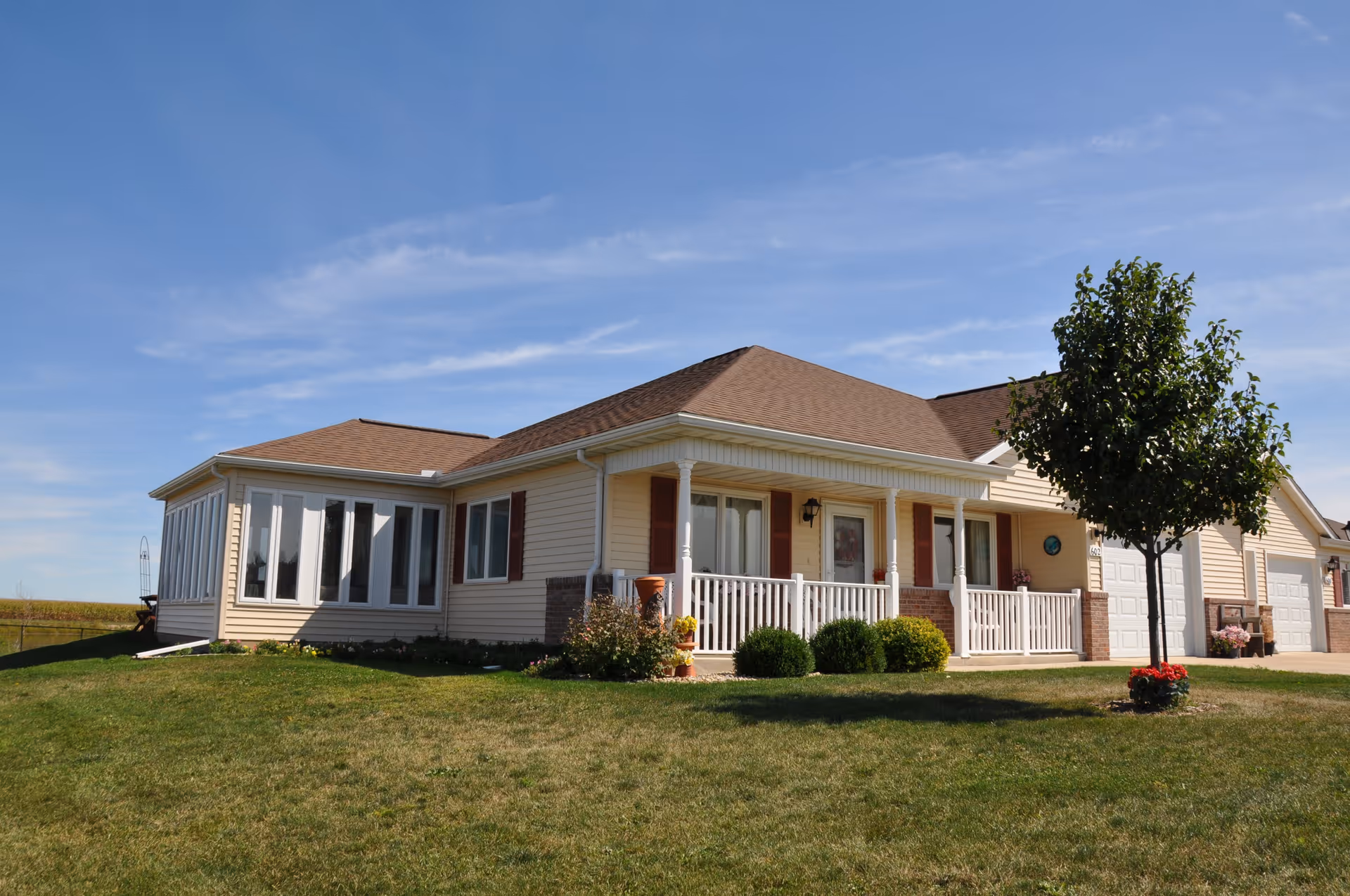 Single-story residential building with beige siding, a brown shingled roof, white porch railing, and a small tree with red flowers in front on a green lawn under a blue sky.