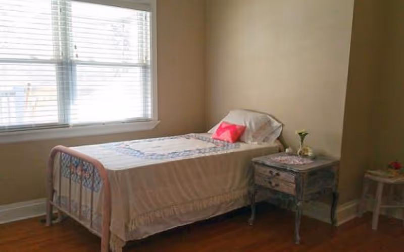 A simple bedroom with a single bed covered with a quilt and a white pillow with a pink cushion. Next to the bed is a small vintage-style nightstand with a flower vase and a doily. The room has beige walls, wooden flooring, and a window with white blinds letting in natural light.