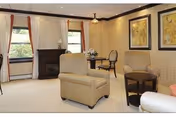 A cozy living room area with beige armchairs, a small round wooden table, a fireplace, two windows with curtains, and framed artwork on the wall.