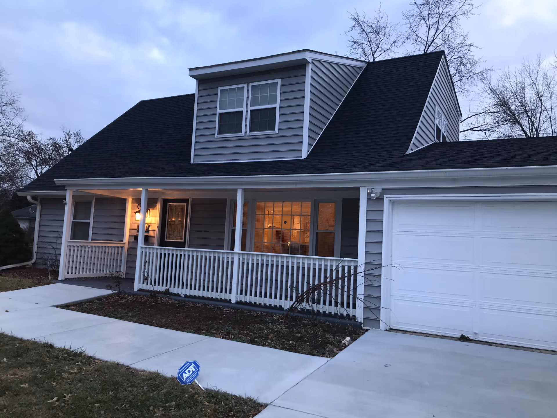 Exterior view of a two-story house with gray siding, a black roof, a white garage door, and a front porch with white railing. The porch light is on, and there is a lit window next to the front door. A concrete walkway leads to the porch, and an ADT security sign is visible on the lawn.