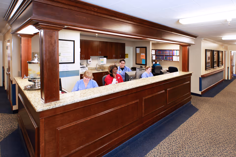 Reception area in Garden City Healthcare Center with a wooden front desk and granite countertop. Four staff members in scrubs are seated behind the desk, working on computers and talking on the phone. The background shows cabinets, office supplies, and a hallway with framed pictures on the wall.