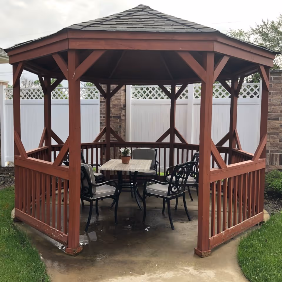 A wooden gazebo with a shingled roof situated on a concrete patio. Inside the gazebo, there is a square table with a small potted plant on top, surrounded by four cushioned chairs with metal frames. The gazebo is located outdoors with a white fence and brick pillars in the background.