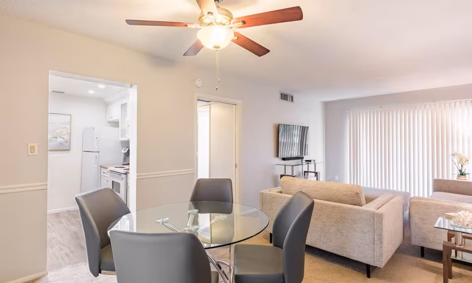 Interior view of a senior living facility showing a dining area with a round glass table and four gray chairs, a living room with beige sofas, a wall-mounted TV, and a sliding glass door with vertical blinds. The kitchen is visible through an open doorway, featuring white cabinets and appliances. A ceiling fan with a light fixture is mounted on the ceiling.