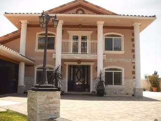 Front exterior view of a two-story house with a tiled roof, four white columns supporting a balcony, large windows, a decorative front door, and a lamp post on a stone pedestal in the driveway.