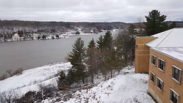 View of a river with a snowy landscape and trees on the riverbank. On the right side, part of a multi-story brick building with several windows is visible. The sky is overcast with gray clouds.