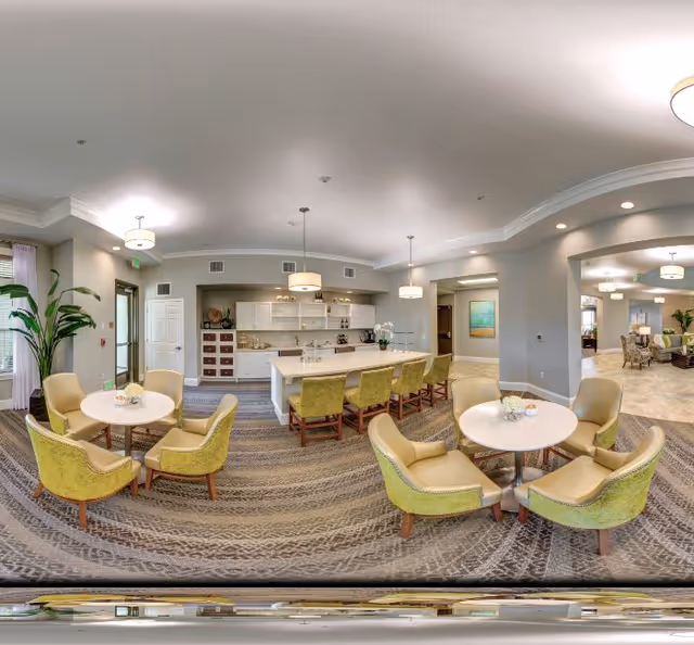 A spacious and well-lit common area in a senior living facility featuring a kitchen with a large island and bar stools, two round tables each surrounded by four green upholstered chairs, and a view into an adjacent living room area with additional seating and decor.