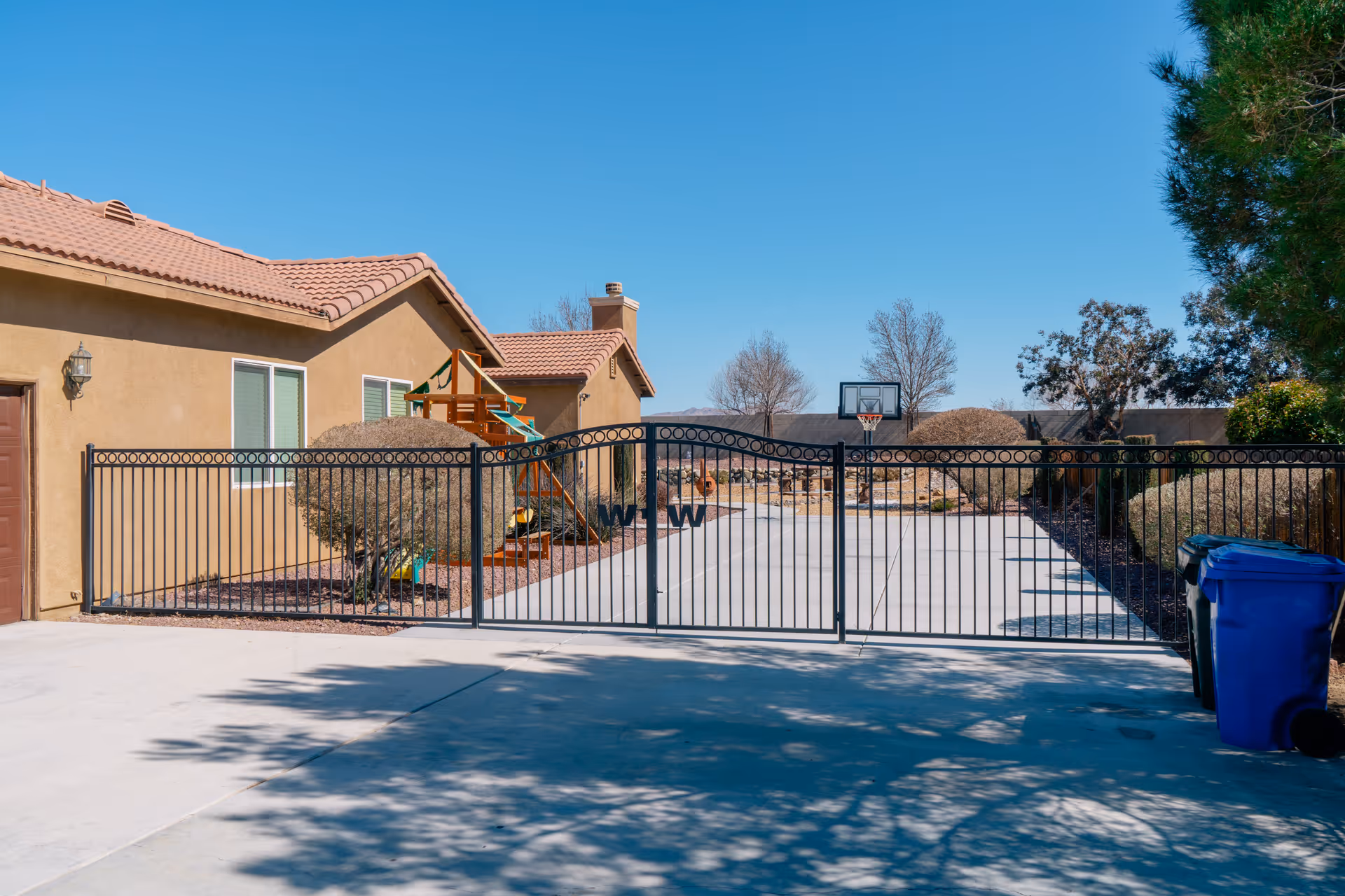 View of a gated driveway leading to a beige stucco house with a tiled roof. There is a basketball hoop at the end of the driveway, some trimmed bushes along the sides, and a blue recycling bin on the right side. The sky is clear and blue.