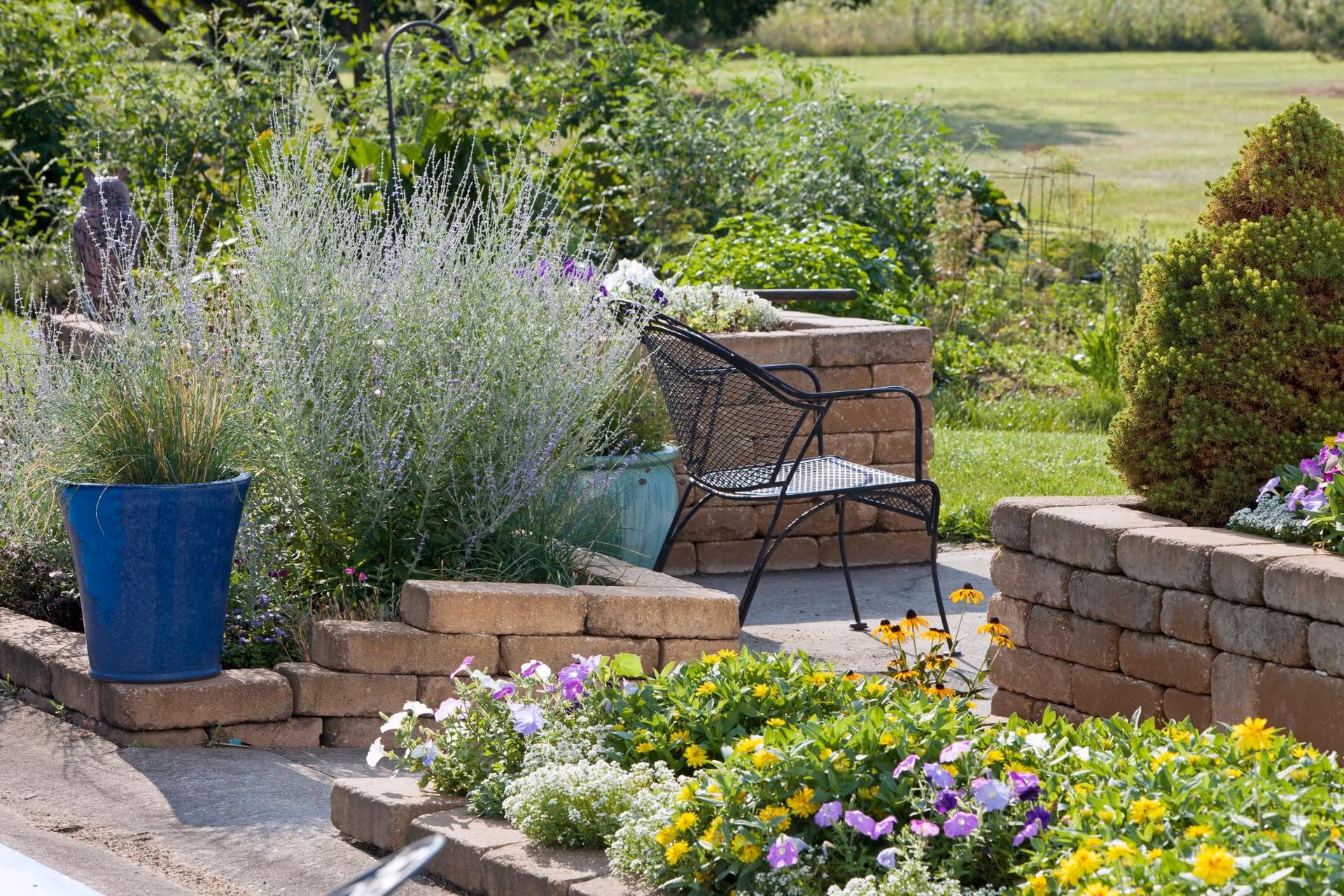A peaceful outdoor garden area with a black metal chair surrounded by raised stone flower beds filled with various colorful flowers and green plants. There is a blue pot with tall grass-like plants and a neatly trimmed bush in the background, with a grassy field beyond the garden.