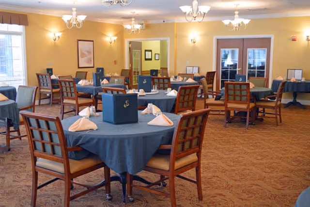 A dining room with multiple round tables covered with blue tablecloths, each set with folded napkins, menus, and salt and pepper shakers. The room has yellow walls, carpeted floors, wooden chairs with cushioned seats, and chandeliers hanging from the ceiling. There are windows on the left side and double doors at the back.