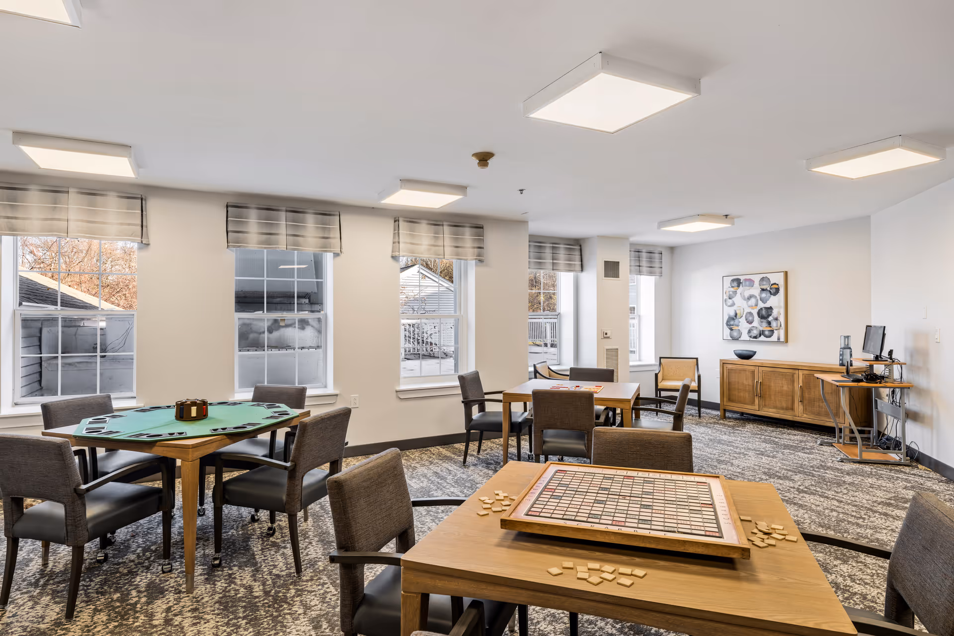A bright and spacious common room with multiple tables and chairs arranged for games. One table has a Scrabble board with tiles scattered around, and another table has a green felt surface with card holders. The room has large windows with striped valances, patterned carpet, modern ceiling lights, a wooden sideboard with decorative items, and a computer workstation in the corner.